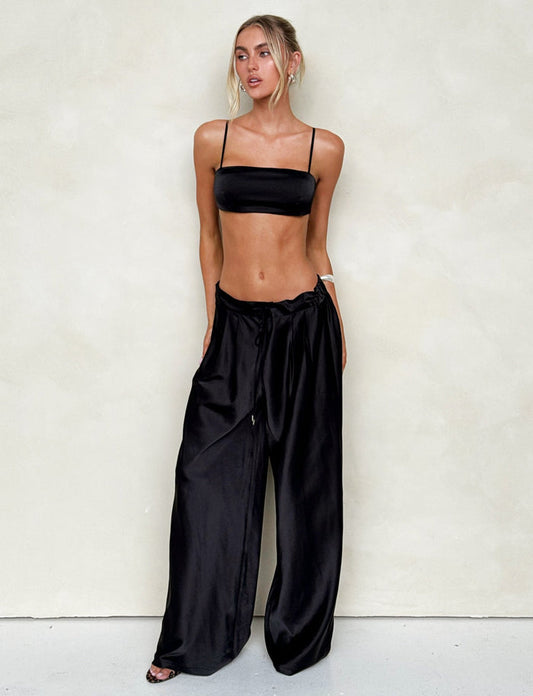 A woman stands against a plain light-colored background, wearing the sleek ALINA TOP - BLACK from TIGERMIST and high-waisted, wide-leg pants. She has blonde hair tied back with loose strands framing her face and is looking slightly to her right.