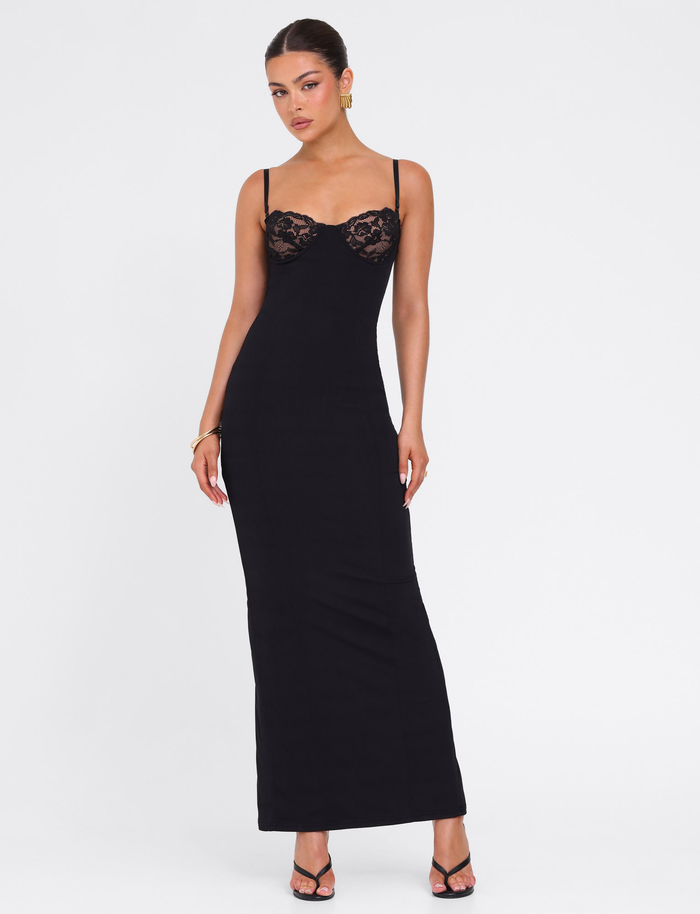 A woman poses against a white background wearing the TIGERMIST ALLURA MAXI DRESS in black, featuring thin straps and lace bra cups. Her hair is tied back, and she accessorizes with earrings, a bracelet, and black heeled sandals.