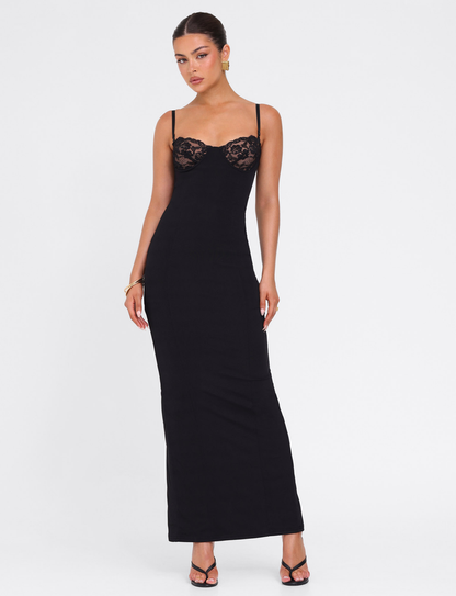 A woman poses against a white background wearing the TIGERMIST ALLURA MAXI DRESS in black, featuring thin straps and lace bra cups. Her hair is tied back, and she accessorizes with earrings, a bracelet, and black heeled sandals.