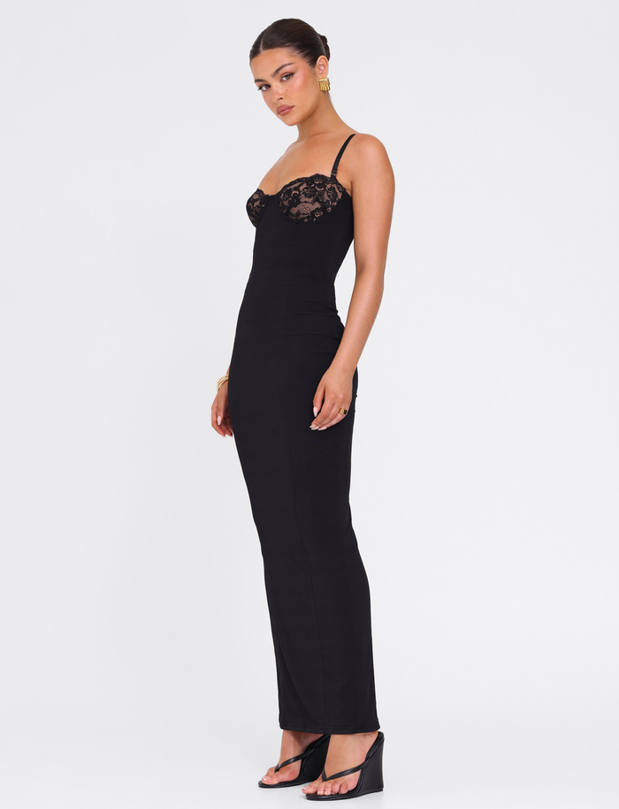 A woman stands sideways wearing the TIGERMIST ALLURA MAXI DRESS in black with lace bra cups, spaghetti straps, and black open-toe heeled sandals. Her hair is up, accessorized with gold earrings and a bracelet.