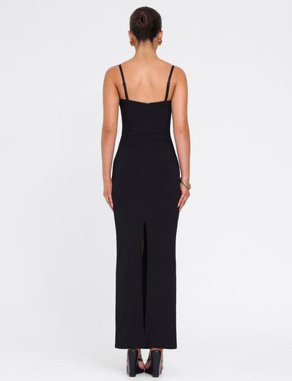 A woman faces away in the TIGERMIST ALLURA MAXI DRESS - BLACK, a fitted bodycon style featuring thin straps, lace bra cups, and a back slit. She pairs it with black heels and a bracelet, her hair in a bun against a plain white background.