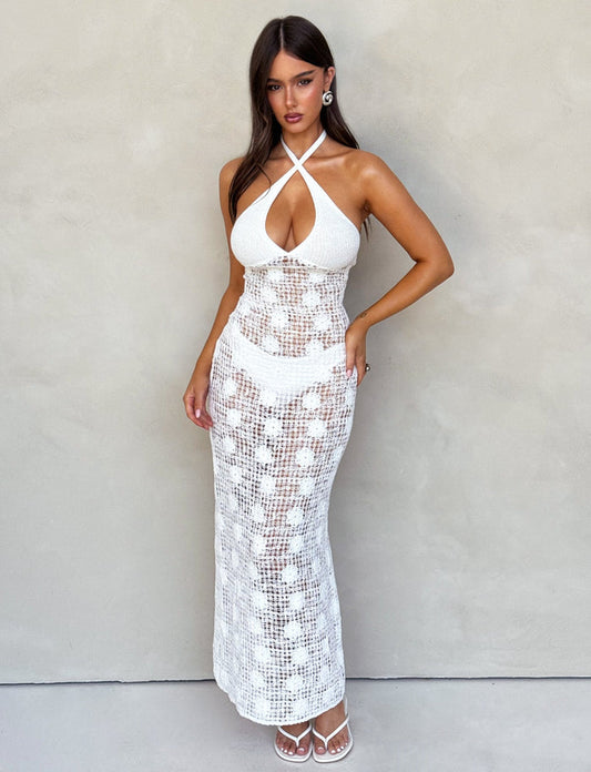 A woman stands against a neutral beige wall, wearing the ARDEEN DRESS - WHITE from TIGERMIST. The dress features a halter-neck style with an open front cut-out and a crochet-like pattern. She has long brown hair and is accessorized with large hoop earrings and white strappy heels, posing with her hand on her hip.