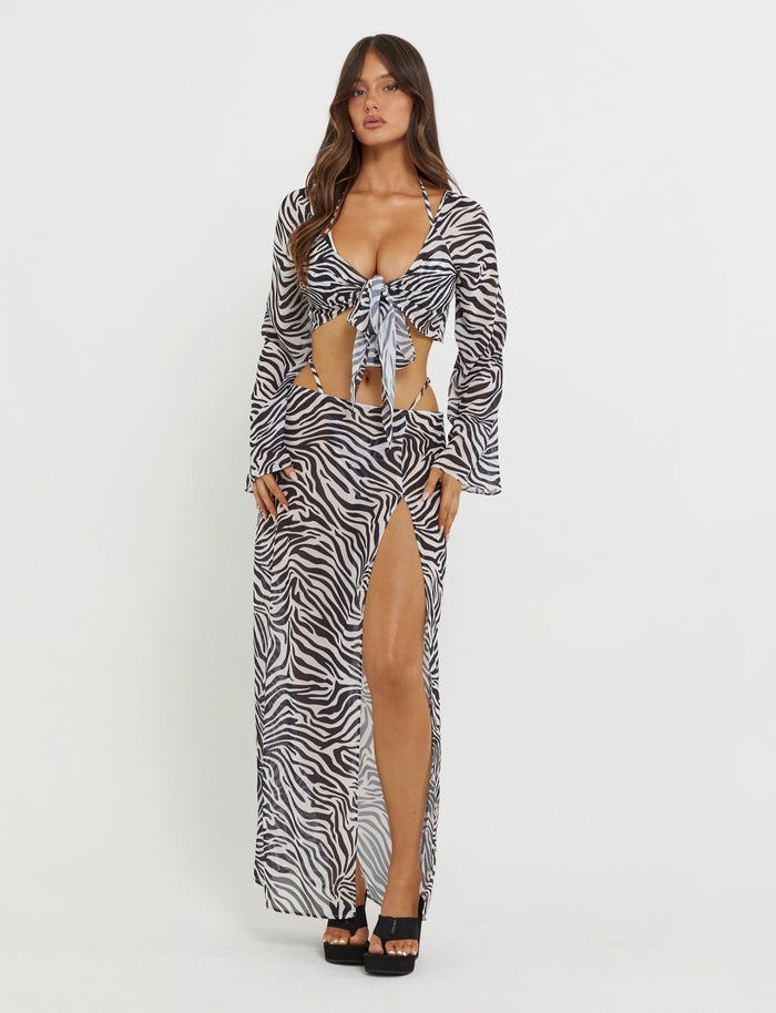 A woman stands against a plain white background, showcasing the TIGERMIST AYESHA TOP - BLACK : ZEBRA. The ensemble includes a Tiger Mist black-and-white zebra print top tied at the front and a long skirt with a high slit. She pairs it with black high-heeled sandals, her long, wavy brown hair cascading down.