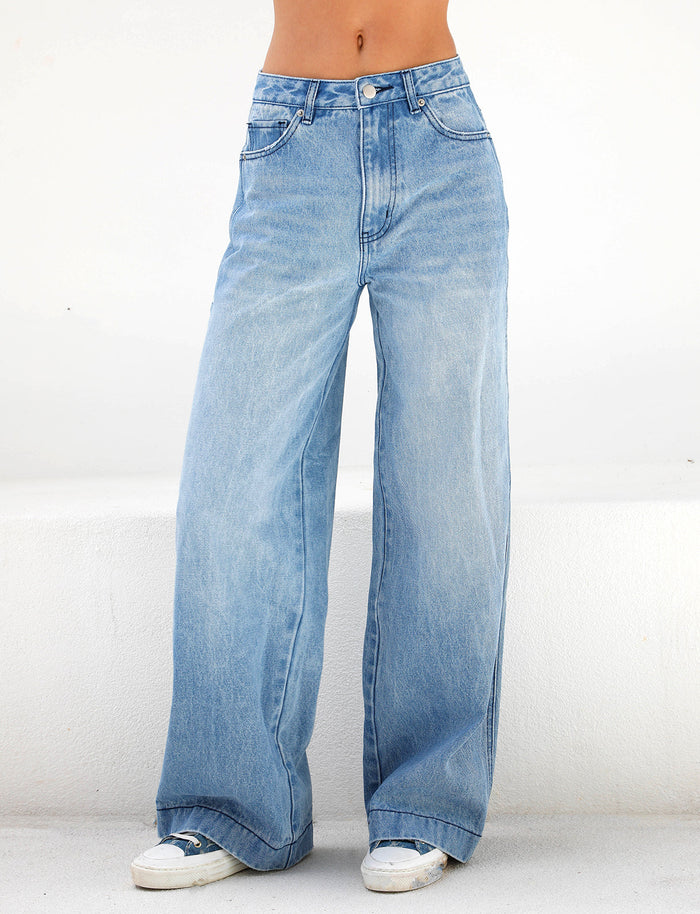 A person stands against a plain white background, wearing the TIGERMIST COHEN JEAN - DENIM, showcasing its wide-leg silhouette in light blue denim. These jeans highlight a high-rise waist and classic five-pocket design, making them a perfect match with crisp white sneakers.