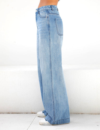 A side view of a person wearing the TIGERMIST COHEN JEAN - DENIM, featuring a high-rise waist and wide-leg cut, paired with white sneakers against a light-colored background. The jeans provide a casual, relaxed fit that flows elegantly to the floor.