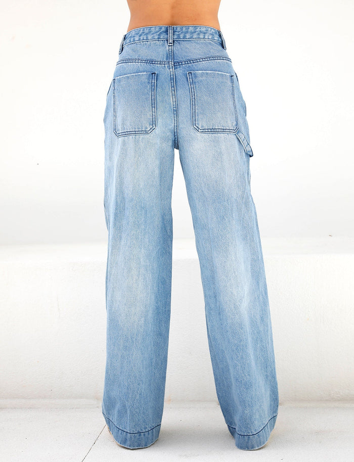 An individual is standing against a plain white background, wearing the TIGERMIST COHEN JEAN - DENIM in a light blue shade. These jeans are designed with large back pockets and a wide-leg silhouette, providing a casual and relaxed fit.