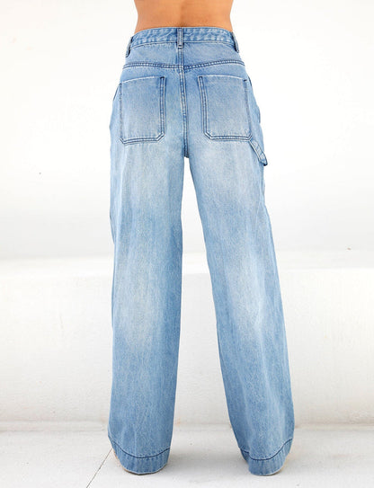 An individual is standing against a plain white background, wearing the TIGERMIST COHEN JEAN - DENIM in a light blue shade. These jeans are designed with large back pockets and a wide-leg silhouette, providing a casual and relaxed fit.