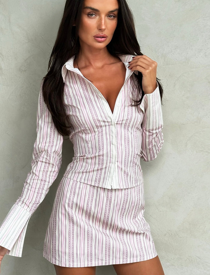 A woman with long, dark hair wears the DARCEE SHIRT - PINK from TIGERMIST along with a matching skirt in pink pinstripe. She stands against a plain textured background, her left hand touching her hair near her collar. She looks confidently to the side.
