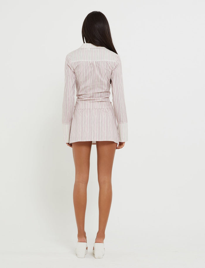 A person with long, dark hair is facing away from the camera, wearing the DARCEE SHIRT in pink pinstripe from TIGERMIST. The light-colored, long-sleeve dress has a fitted waist and ends at mid-thigh. They are also wearing white heeled shoes. The background is plain white.