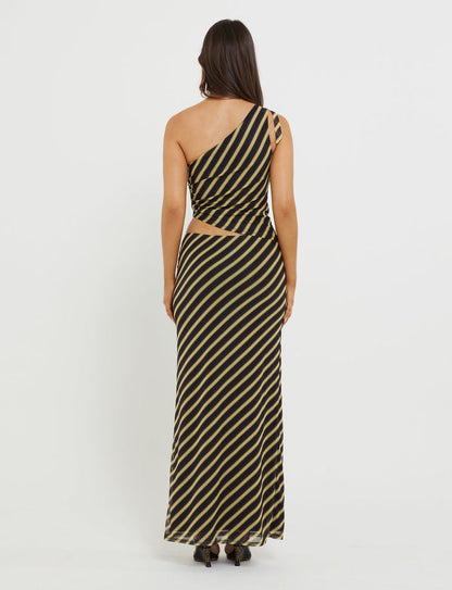 A person with long, dark hair stands facing away, wearing the DEANNA MAXI SKIRT - MULTI : STRIPE : BROWN STRIPE from TIGERMIST. The skirt features diagonal black and beige stripes and a cut-out detail on the right side at the waist. The person is also wearing black, open-toed heels.