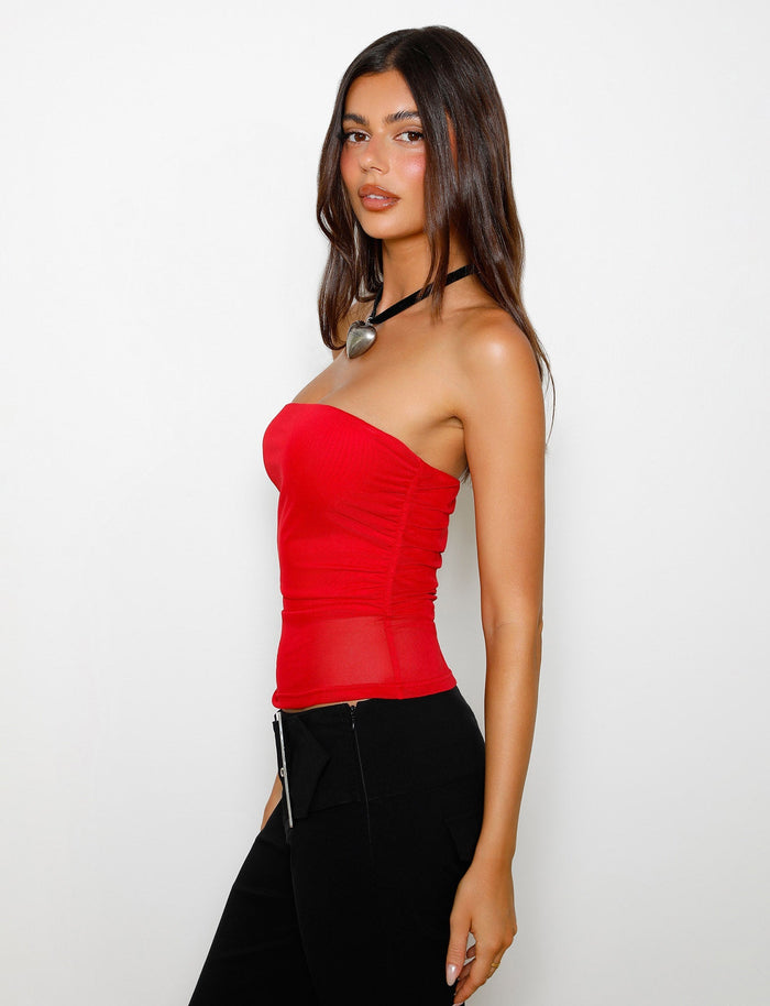 A person with long dark hair is wearing the HARRIETT TUBE TOP - RED from TIGERMIST along with black pants. They are standing against a plain white background, looking at the camera with a neutral expression. In addition, they are accessorized with a thick black choker featuring a large pendant.