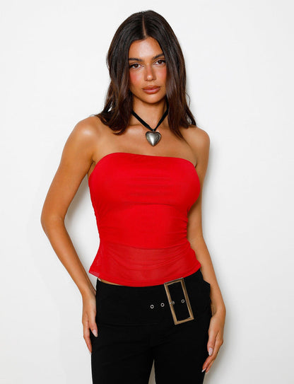 A person with long dark hair is wearing the TIGERMIST HARRIETT TUBE TOP in red and black pants with a large rectangular belt buckle. They are also adorned with a large heart-shaped necklace against a plain white background.