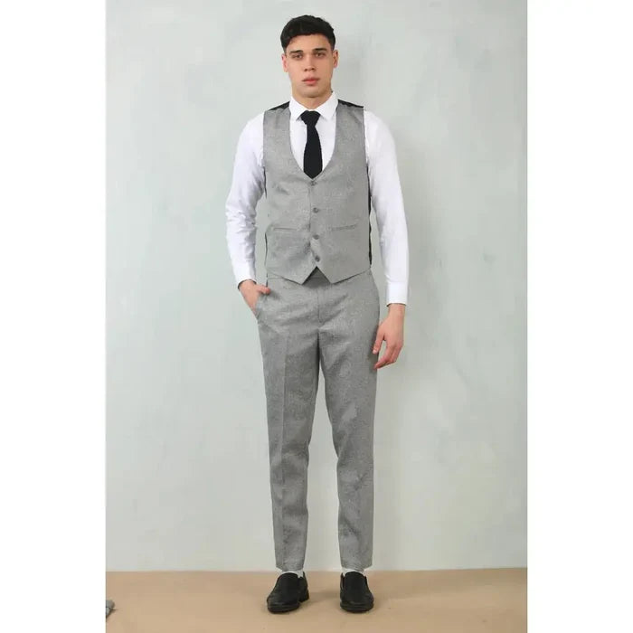 Back view of a man in a grey tailored fit wedding 3-piece suit with a white shirt, black shoes, and a black vest, standing against a plain wall.