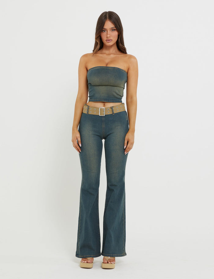 A woman stands against a plain white background, wearing the TIGERMIST ISABEL JEAN - BLUE denim tube top and matching high-waisted, flared jeans. She has long, straight hair and is accessorized with a wide beige belt featuring metallic accents and platform sandals.