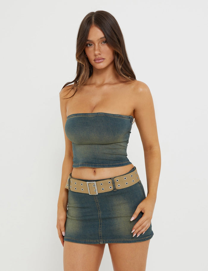 A woman stands against a plain white background, wearing a chic ISABEL TUBE TOP - BLUE : DENIM BLUE from TIGERMIST, paired with a matching denim mini skirt. She accessorizes with a beige belt featuring silver eyelets. Her hair is down, and she maintains a neutral expression.