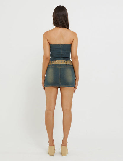 A woman with long, dark hair stands facing away from the camera. She is wearing the ISABEL TUBE TOP - BLUE : DENIM BLUE by TIGERMIST, paired with a wide, studded belt and beige wedge shoes. The background is white and plain.