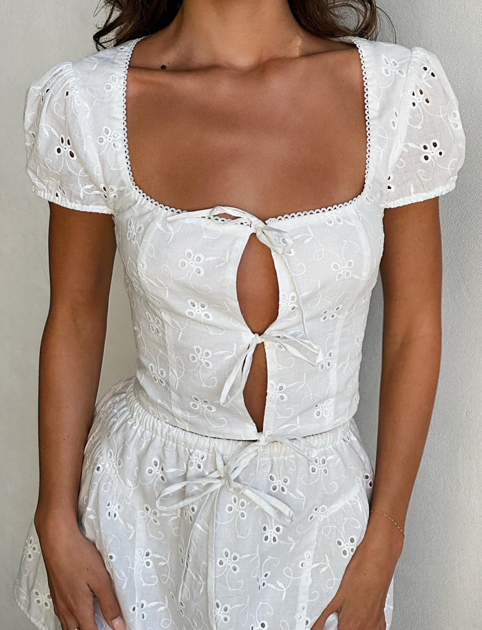 A person is wearing the Kayleigh Top in white from TigerMist. This top is eyelet-embroidered with short puff sleeves and a square neckline, featuring keyhole cutouts tied with bows down the front. Their arms are relaxed by their sides, and their skin tone is medium tan.