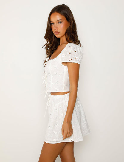 A person with long brown hair stands against a plain white background, wearing the TIGERMIST KAYLEIGH TOP - WHITE, featuring an eyelet pattern with short puff sleeves and a matching skirt. They are facing slightly to the left, with a neutral expression.