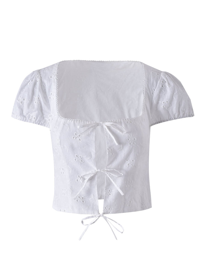 The TIGERMIST Kayleigh Top in white features a square neckline, short sleeves, and eyelet embroidery. It includes three center-front tie closures and showcases a delicate floral pattern on the fabric. This slightly cropped blouse effortlessly blends elegance with a touch of playful charm.