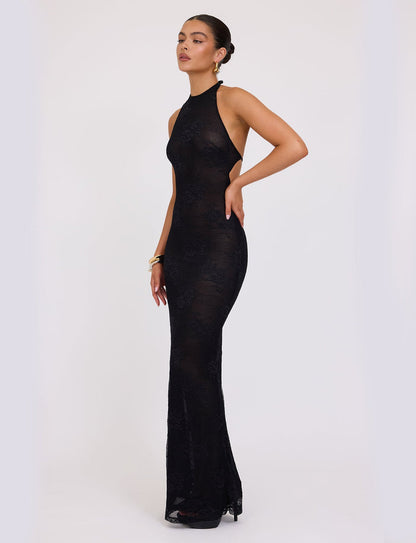A woman is wearing the sophisticated LUCIA DRESS - BLACK : LACE from TIGERMIST, which features a high neck and sleeveless design. The form-fitting gown has a sheer appearance with an elegant, subtle floral pattern. She is posing gracefully with one hand on her hip and the other arm slightly bent, accessorized with hoop earrings and a bracelet.