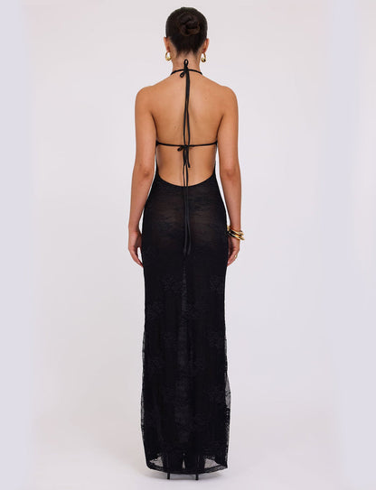 A woman stands with her back to the camera, wearing the LUCIA DRESS - BLACK : LACE from TIGERMIST. The black halter-neck gown features an open back secured with thin straps, and she has styled her hair in a bun. Her ensemble is complemented by gold hoop earrings and gold bangles. The floor-length dress is slightly transparent.