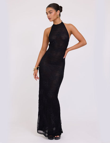 A woman is posing in a full-length, form-fitting LUCIA DRESS - BLACK : LACE from TIGERMIST with a halter neckline. She accessorizes with a gold bracelet and hoop earrings, has her hair styled up in a bun, and stands against a plain white background.