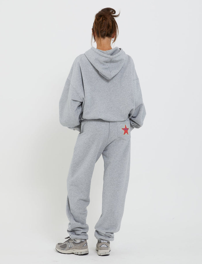 A person stands facing away from the camera, wearing a light gray LUMEN HOODIE by TIGERMIST and matching sweatpants with a red star on the back pocket. They also have gray sneakers on, and their hair is tied up in a messy bun. The background is plain white.