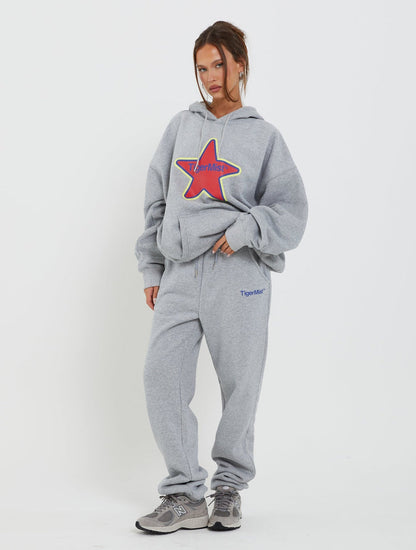 A woman is posing against a white background, donning a light gray LUMEN HOODIE by TIGERMIST with a colorful star graphic, matching gray sweatpants featuring "Tiger Mist" on one leg, and gray athletic shoes. Her hair is tied back, and she has a relaxed stance.