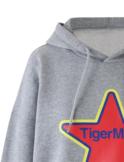 A close-up view of the LUMEN HOODIE - GREY by TIGERMIST, showcasing its light gray color and drawstring feature. The hoodie displays a prominent, colorful graphic of a red star with yellow and blue outlines on the front. Inside the star, "Tiger Mist" is partially visible in bold blue letters.