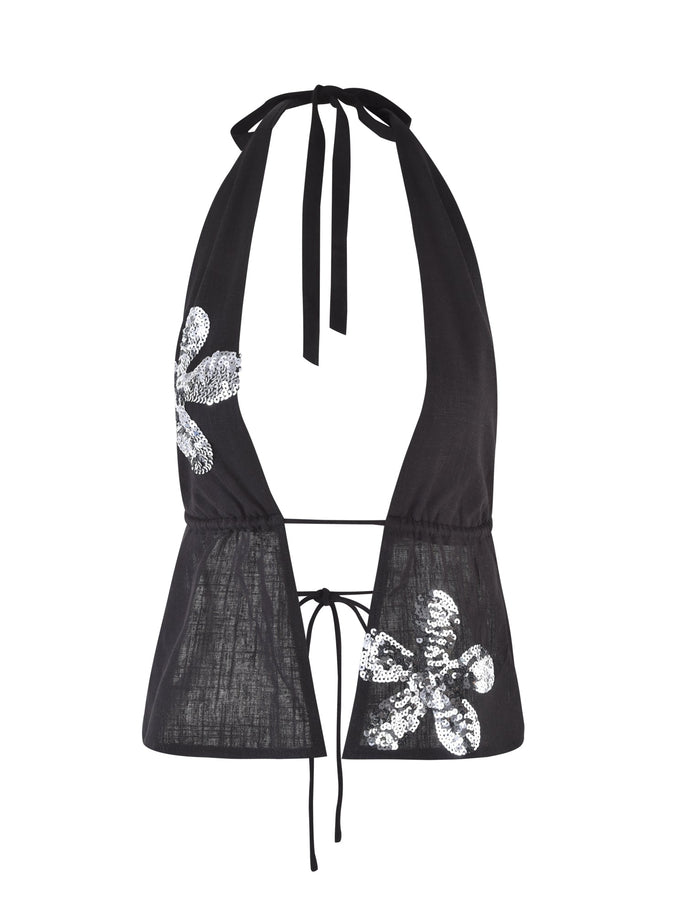 The MAISHA TOP from TIGERMIST is a black halter top with a deep V-neckline, featuring tie straps at the neck and back. It is adorned with sequined floral appliques on both sides, adding a touch of sparkle and elegance to the simple design.
