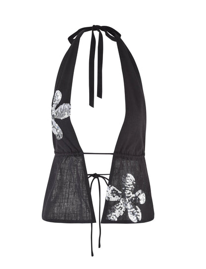 The MAISHA TOP from TIGERMIST is a black halter top with a deep V-neckline, featuring tie straps at the neck and back. It is adorned with sequined floral appliques on both sides, adding a touch of sparkle and elegance to the simple design.