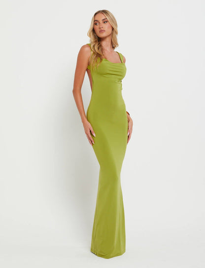 A woman stands against a plain white background, wearing the MORANA DRESS - GREEN : PASTEL GREEN from TIGERMIST. Her long blonde hair cascades in loose waves over her shoulders. She gazes off to the side with one hand resting on her hip and the other hanging by her side.