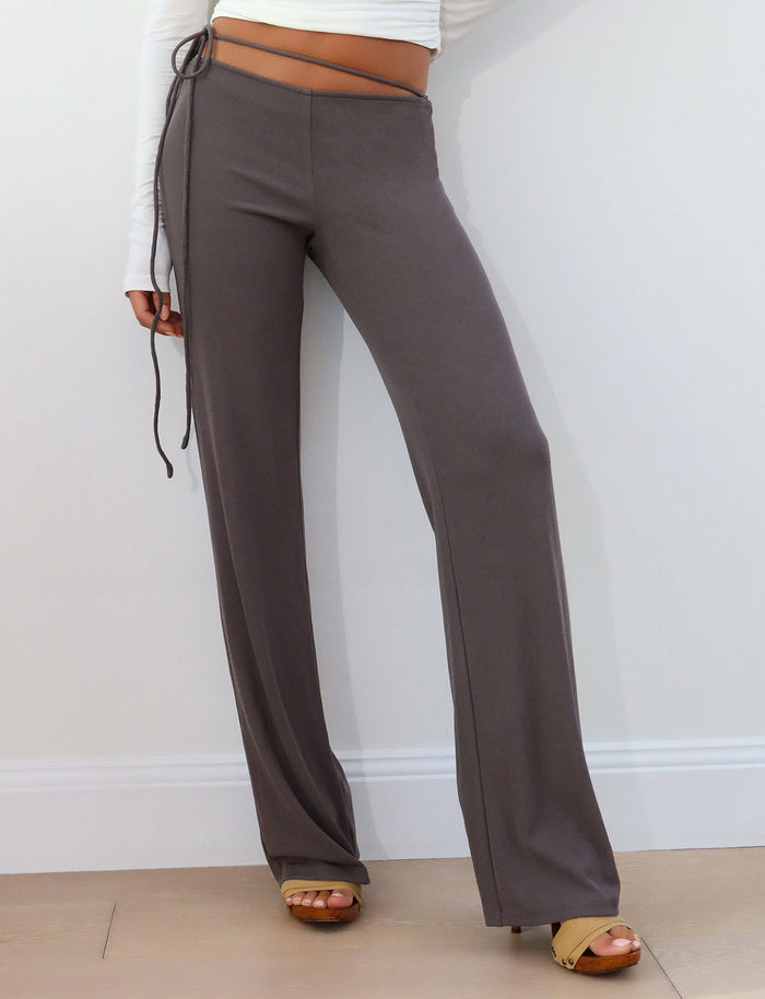 A person stands against a white wall, wearing the REY PANT - GREY : CHARCOAL from TIGERMIST. They pair the high-waisted, wide-leg charcoal trousers with a white long-sleeve crop top from TIGERMIST. Posing with their left hand on their hip and right arm crossing their body, they complete the look with light brown open-toe heels.