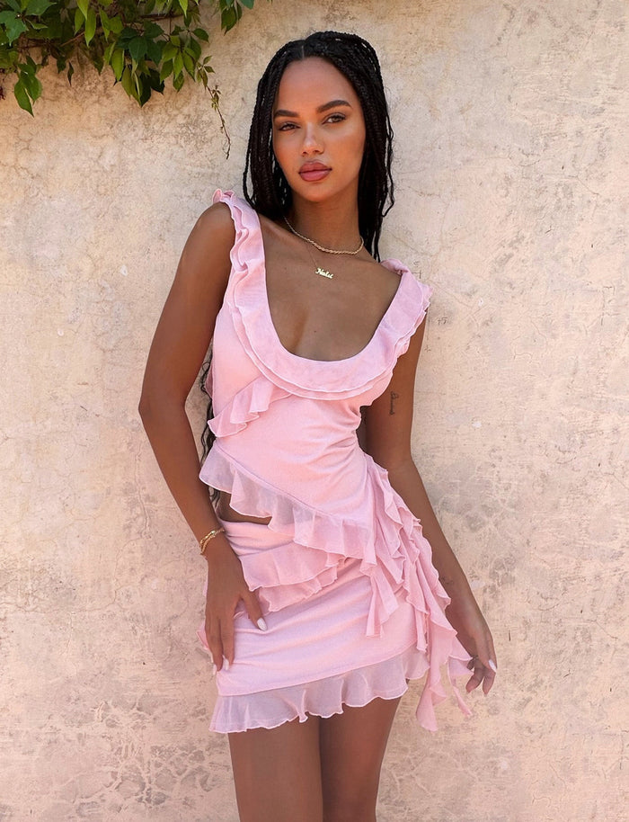 A woman stands against a textured beige wall, wearing the Baby Pink Stormi Top from Tigermist. Her hair is styled in long braids, and she's accessorized with a gold necklace and bracelet. She poses confidently, resting one hand on her hip and the other beside her. Greenery is visible above her.