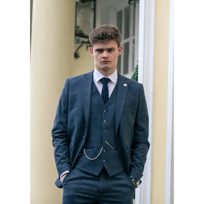 Young man in dark gray tailored suit with matching trousers, white shirt, and black shoes, standing outdoors.