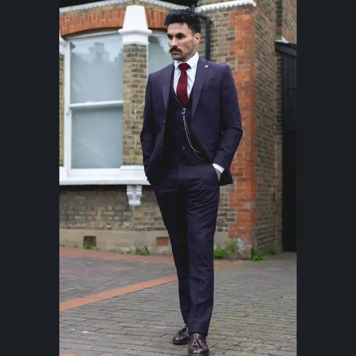 Man wearing a stylish dark plum wine check 3-piece suit with white shirt, red tie, and pocket square, standing outdoors in urban setting.