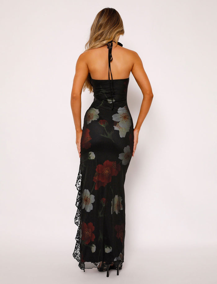 A woman with long blonde hair wearing the TIGERMIST THEO MAXI DRESS - RED : MACRO FLORAL, featuring a stunning black halter-neck, floor-length design adorned with a large red, white, and green floral print. She stands with her back to the camera, highlighting the dress's backless style and intricate lace detailing along the sides.
