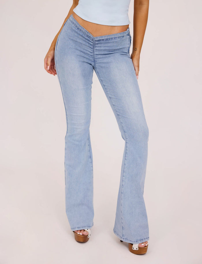 A person wearing the light blue TIJANA JEAN - BLUE from TIGERMIST, which are high-waisted flared jeans featuring a V-shaped waistband, paired with a crop top. The person is standing with one hand resting on their thigh and the other hanging by their side against a plain white background.
