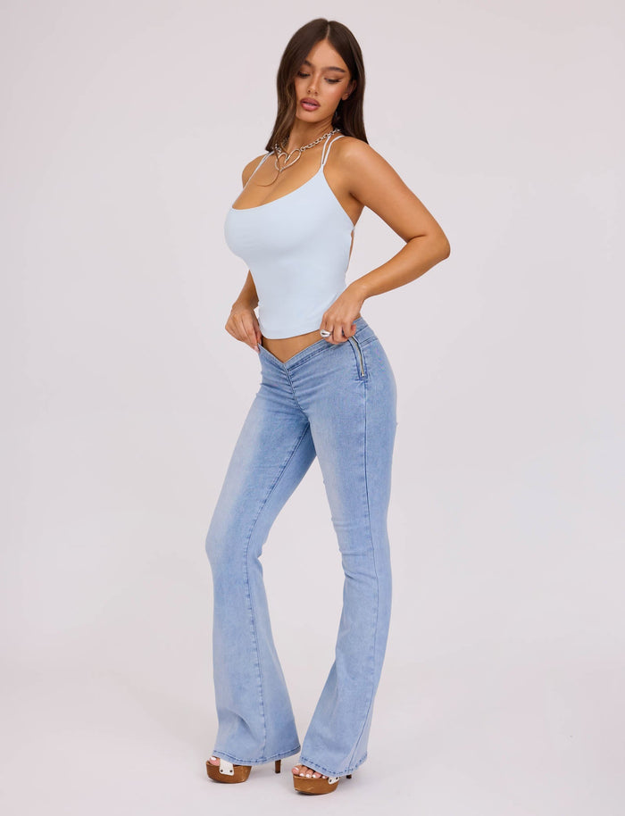 A woman stands against a plain background, wearing a light blue spaghetti strap top and the TIGERMIST TIJANA JEAN - BLUE : DENIM BLUE. She has long brown hair and is posing with one hand on her hip and the other touching the waistband of her jeans. She completes her outfit with white open-toed heels.