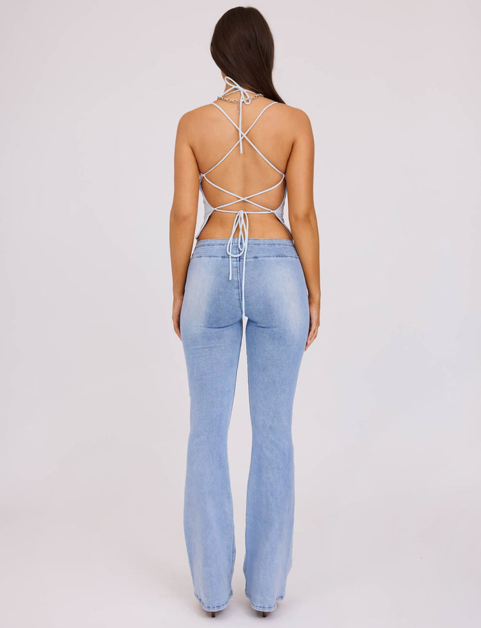 A person with long dark hair stands facing away, wearing a white strappy backless top from TIGERMIST and light blue high-waisted TIJANA JEAN - BLUE : DENIM BLUE from TIGERMIST. The background is plain white.