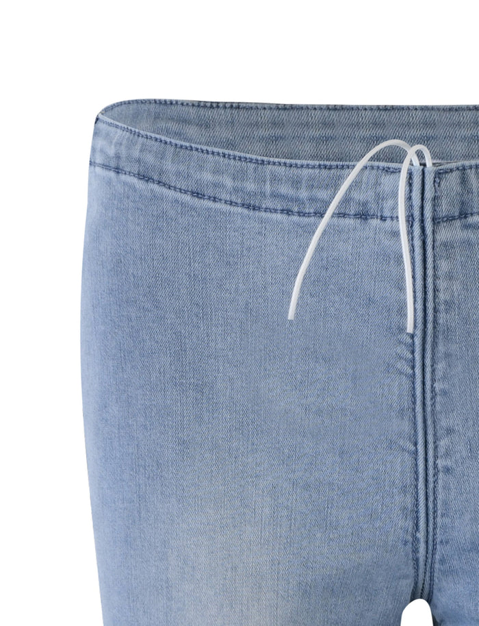 Close-up of the TIJANA JEAN - BLUE: DENIM BLUE from TIGERMIST. The top part of the light blue denim jeans is visible, featuring a drawstring waist extending from the center of the waistband. The fabric appears slightly textured.