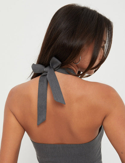A person with long, dark hair tied in a low ponytail is shown from the back. They are wearing the TRISTAN VEST - GREY by TIGERMIST, which ties into a bow at the nape of the neck. The background is plain white.