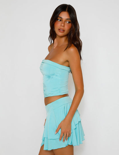 A woman with long, dark hair is dressed in the TIGERMIST VERANIQUE TOP - BLUE and a matching pleated skirt. She stands sideways, looking over her shoulder against a plain white background.